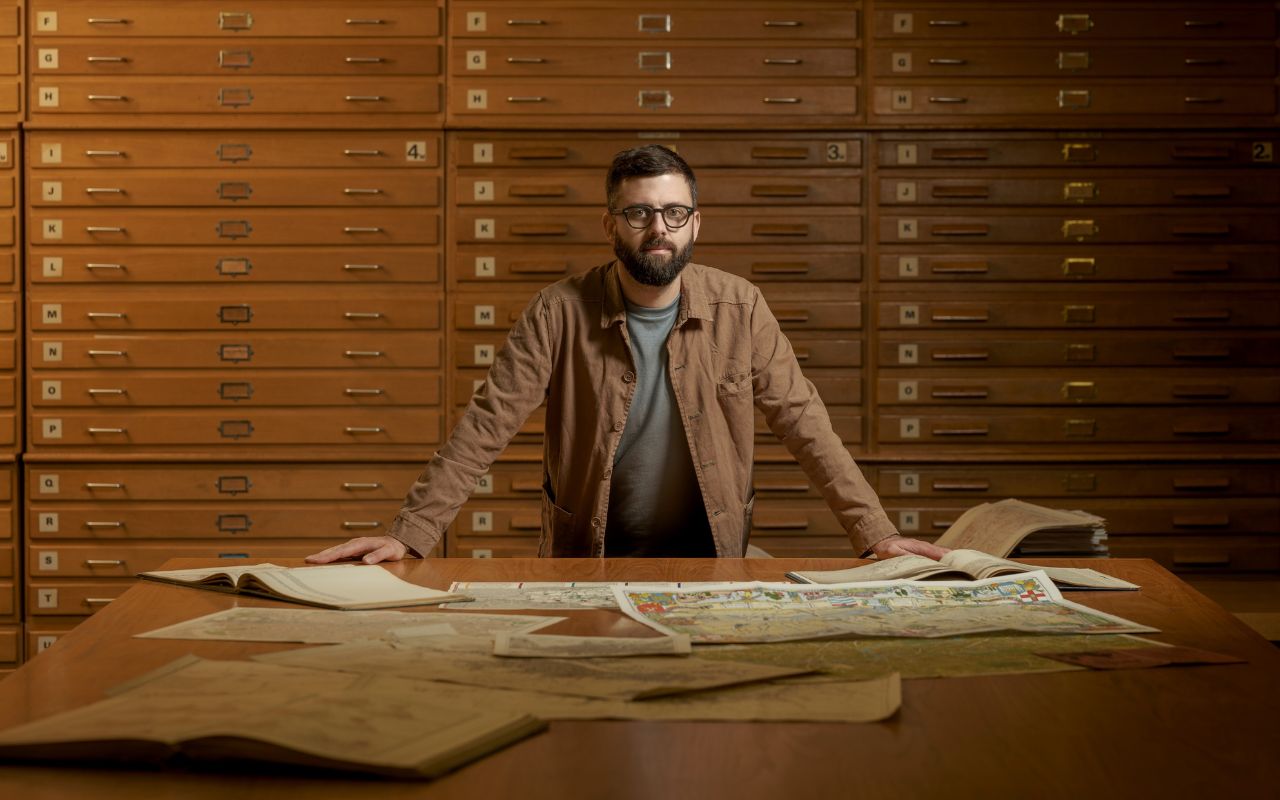 Professor James Cheshire stood in front of a large table with open books and large maps. Behind him are a set of floor to ceiling archive drawers archive drawers in a soft mustard colour.