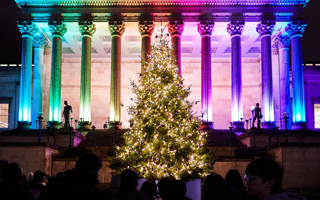 UCL's Wilkins building illuminated in rainbow colours at night, with a brightly lit Christmas tree in front and a crowd of people gathered around it taking photographs.